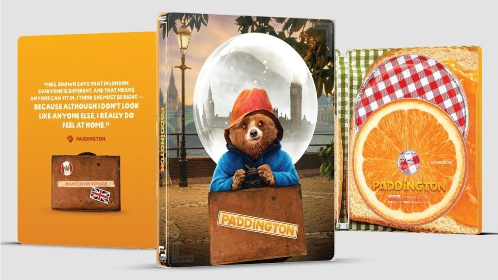 Steelbook case featuring Paddington Bear in a blue coat and red hat, peeking out from a suitcase with a backdrop of London landmarks. Includes a quote about feeling at home and visual elements of Paddington's adventure.